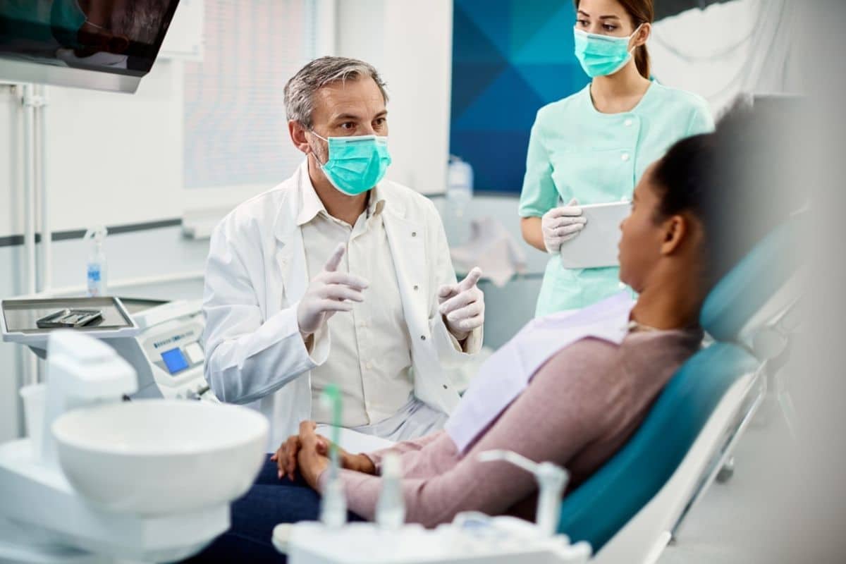 Dentist in mask examining patient in dental chair with assistant present in modern clinic.
