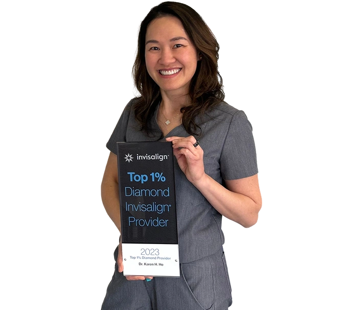 A smiling woman in grey dental scrubs holding a black-and-white Invisalign award plaque against a solid teal background. The award reads "invisalign Top 1% Diamond Invisalign Provider" at the top, and "2023 Top 1% Diamond Provider Dr. Karen H. Ho" at the bottom.